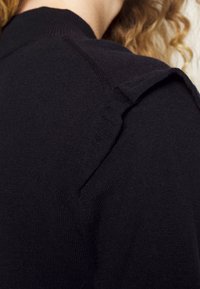 Zbliżenie na ramię osoby noszącej czarny sweter z dzianiny z małym materiałowym klapą na szwie ramienia.