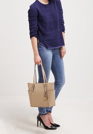 Kvinde i en marineblå sweater og jeans bærer en beige tote bag, mens hun står i høje hæle mod en neutral baggrund.
