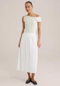 Ljusgrön off-shoulder topp i kombination med en vit maxiklänning och mörka platta skor. Outfitten har mjuka, texturerade tyger och en avslappnad passform.
