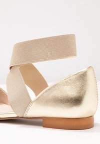 Sandal plano con una punta y un talón de cuero dorado brillante, con una amplia correa elástica beige cruzando la parte superior. Acabado texturizado, diseño minimalista.