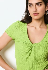 Grünes strukturiertes Oberteil mit kurzenÄrmeln, das einen tiefen V-Ausschnitt und ein Twist-Detail vorne aufweist. Das Material hat ein zerknittertes Aussehen.