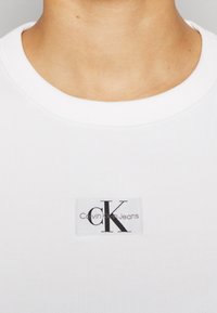 T-shirt bianco a righe con etichetta quadrata con "CK" e "Calvin Klein Jeans" in lettere nere nella zona del petto.