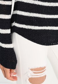 Suéter de punto en azul marino con rayas horizontales blancas, con una trama suelta y mangas largas ligeramente acampanadas; combinado con jeans blancos rasgados.