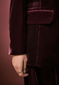 Burgundy velvet blazer with a pocket detail; smooth texture and tailored fit, paired with a hand wearing a silver ring.
