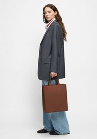 Brązowa torba typu tote ze skóry o prostokątnym kształcie i dwoma górnymi uchwytami. Noszona z szarą marynarką w prążki i szerokimi dżinsami.