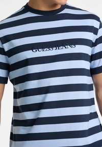 T-shirt rayé avec des rayures horizontales alternées bleu foncé et bleu clair, en coton, col rond et logo "GUESS JEANS" brodé.