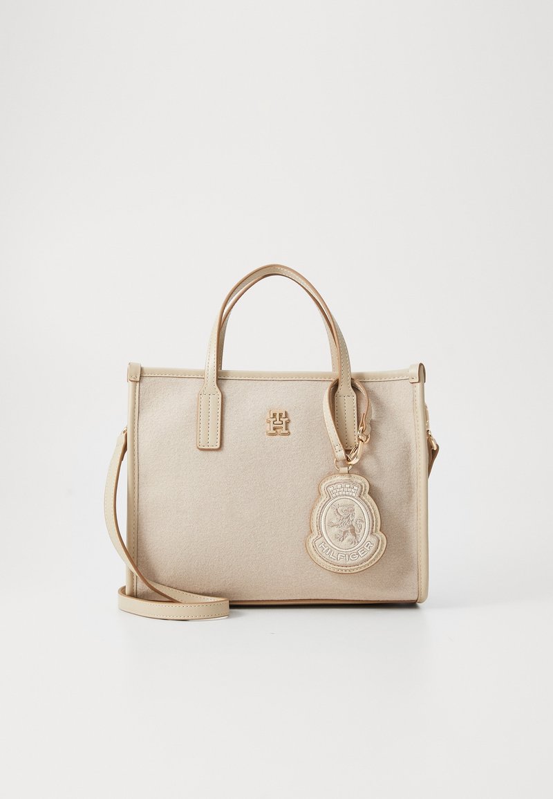 Sac à main rectangulaire beige avec deux poignées, une bandoulière amovible et un patch logo rond suspendu sur le côté avant droit.