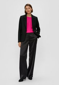 Schwarze, maßgeschneiderte Blazer über einer rosa plissierten Bluse, kombiniert mit hoch taillierten, fast schwarzen Satinhosen und spitzen schwarzen Schuhen.