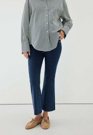 Persona que lleva una camisa de botones a rayas negras y blancas, vaqueros azul oscuro de corte recto y mocasines de ante color tostado. Una mano en el bolsillo.