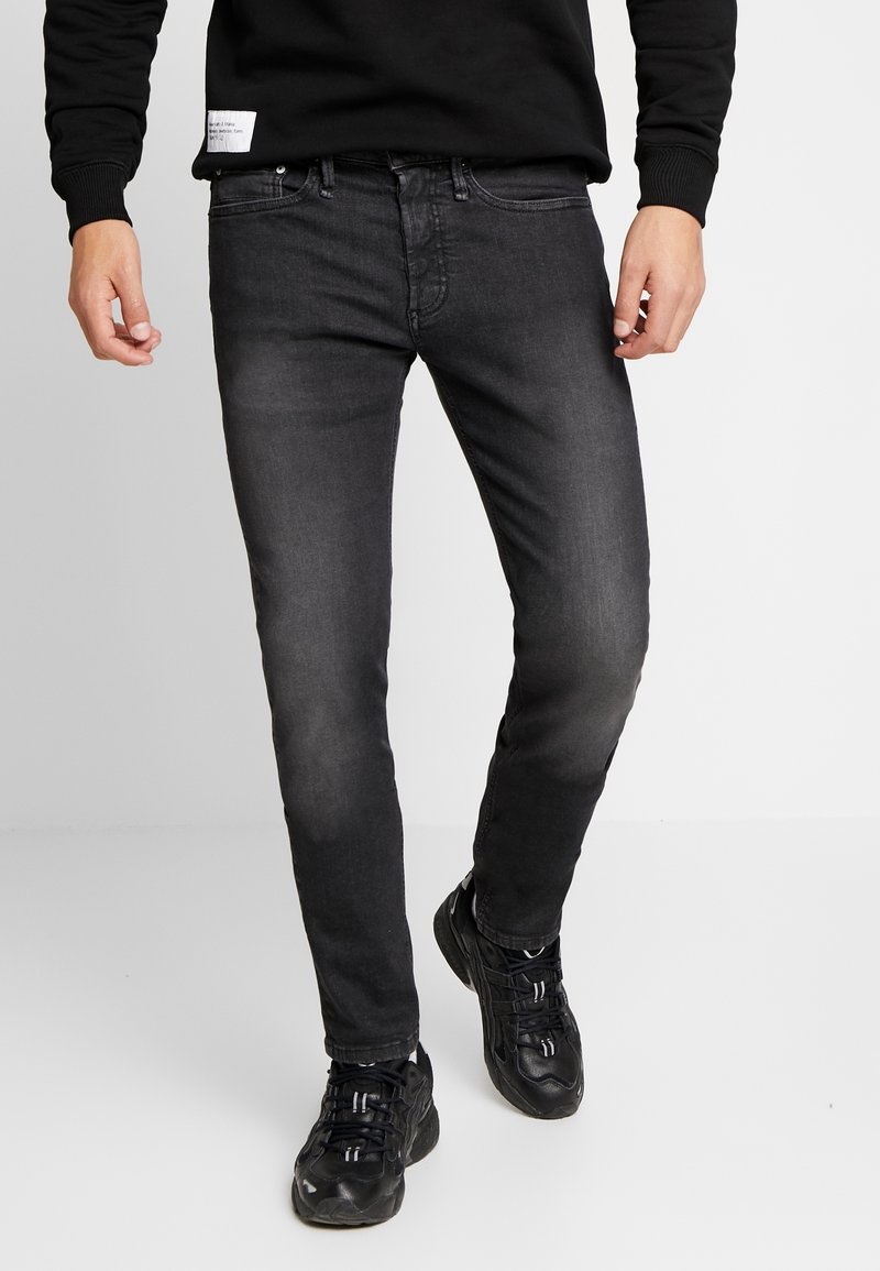 Homme portant un jean noir délavé slim, des baskets noires et un sweat-shirt noir, debout devant un fond clair uni.