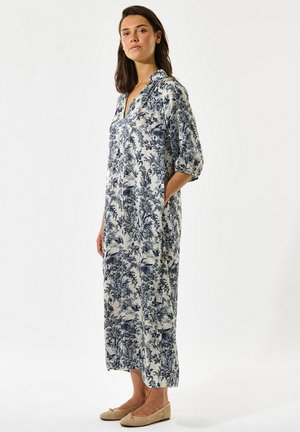 Žena stojaca v dlhej, voľnej bielej sukni s modrými kvetinovými a vtáčími vzormi, s tromi štvrťovými rukávmi a béžovými plochými topánkami.