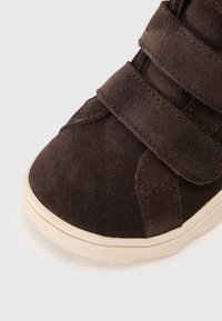 Zapato de ante marrón con punta redonda, que cuenta con dos correas de velcro para el cierre y una suela de goma texturizada color crema.