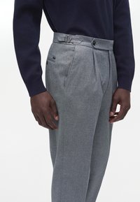 Homme portant un pantalon plissé gris avec boucle de réglage latérale et un haut sombre à manches longues, vu de la taille jusqu'au milieu de la cuisse.