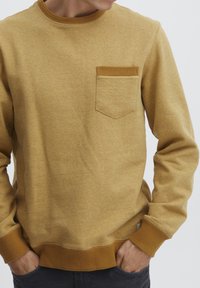 Sudadera amarilla mostaza con cuello redondo, puños acanalados y un pequeño bolsillo en el pecho. Fabricada en un tejido suave y texturizado.