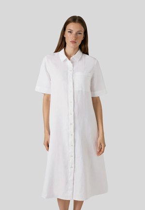 Kvinde iført en hvid, kortærmet midi-kjole med knapper, krave og brystlomme, stående mod en ensartet lysegrå baggrund.