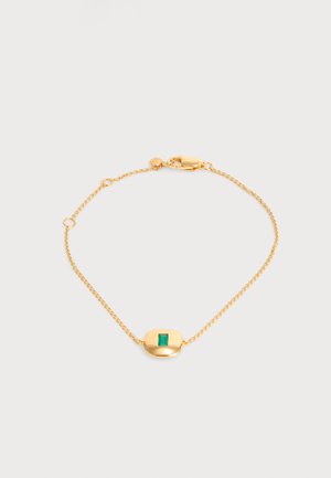 Delicada pulsera de cadena de oro con un charm ovalado de oro que presenta una pequeña piedra preciosa verde rectangular en el centro sobre un fondo claro.