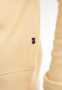 Sweatshirt giallo chiaro in cotone morbido, con un piccolo etichetta nera con logo bianco sulla cucitura laterale. Texture morbida e maniche aderenti.