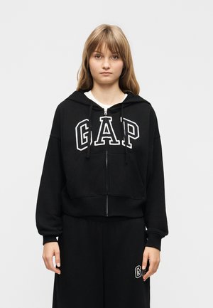 Young woman wearing black zip-up hoodie and pants with "GAP" logo, standing against plain light grey background, expression neutral.