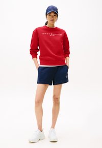 Rød sweatshirt med hvid "TOMMY HILFIGER" logo, parret med marineblå shorts og hvide sneakers. Modellen bærer en marineblå cap.