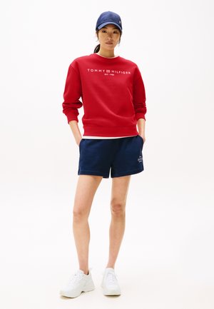 Felpa rossa con logo bianco "TOMMY HILFIGER", abbinata a pantaloni corti blu navy e sneakers bianche. Il modello indossa un cappellino blu navy.
