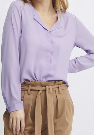Femme portant un chemisier lavande à manches longues rentré dans un pantalon camel taille haute avec une ceinture en tissu nouée.