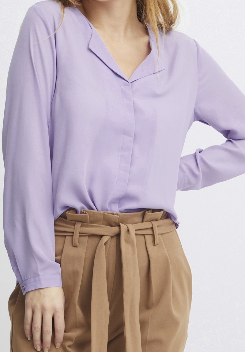 Femme portant un chemisier lavande à manches longues rentré dans un pantalon camel taille haute avec une ceinture en tissu nouée.