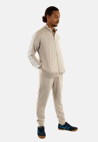 Giacca da track beige con zip e strisce bianche sulle maniche, abbinata a pantaloni tapered coordinati. Sneakers nere e blu completano l'outfit.