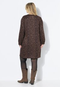 Bruin dierenprint jurk met lange mouwen, een relaxed fit en een keyhole detail op de rug. G gedragen met bruine cowboylaarzen en donkere panty’s.