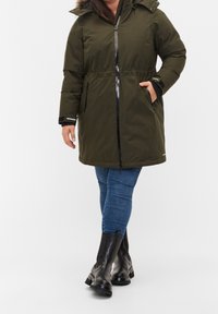 Parka verde oliva, de largo hasta la rodilla, con cierre de cremallera, cintura ajustable con cordón y capucha forrada de piel de pelo. Presenta dos bolsillos laterales y detalles en negro.