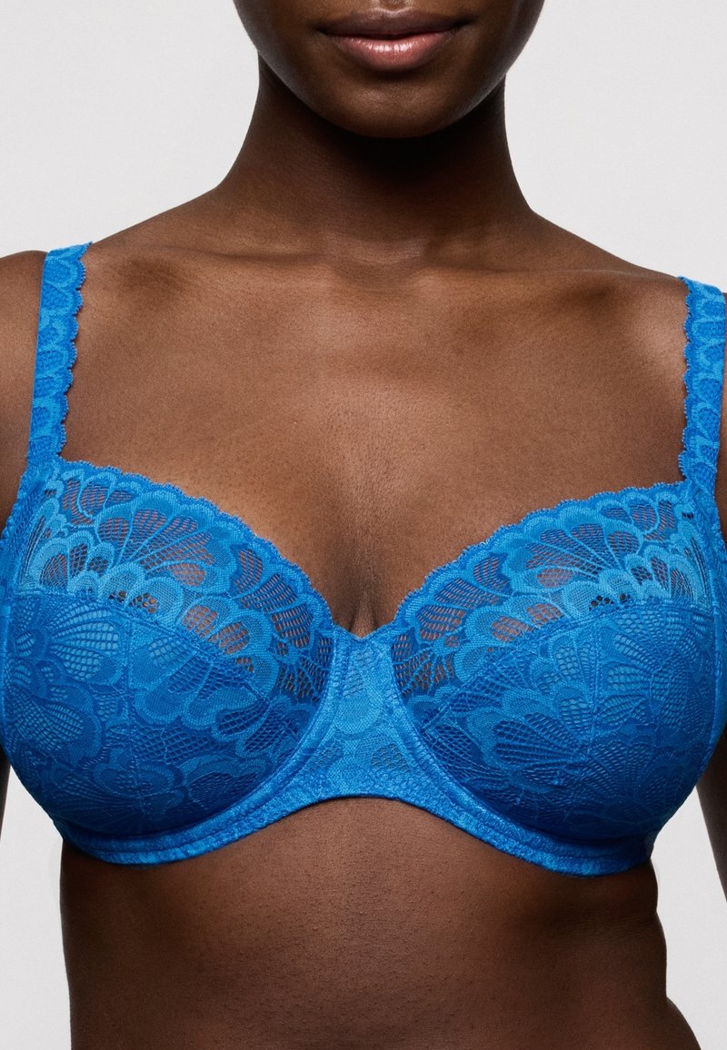 Femme portant un soutien-gorge à armatures en dentelle bleu vif avec des motifs floraux, visible des épaules jusqu'au milieu du torse sur un fond uni.