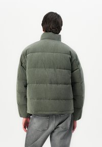 Person seen from behind wearing a green corduroy puffer jacket and gray jeans against a white background.