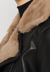 Zwarte leren jas met een kraag van beige kunstbont. Voorzien van een ritssluiting en een gladde textuur, waarbij het contrast tussen de materialen benadrukt wordt.