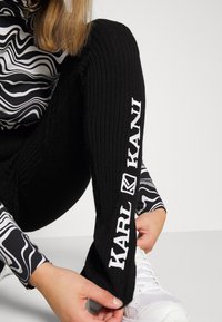 Black ribbed leggings with white embroidered "KARL KANI" branding on the left leg, paired with a black and white patterned long-sleeve top.