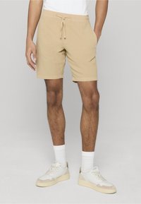 Shorts en coton beige avec une taille élastique, une fermeture à cordon et des poches latérales. Associés à des baskets blanches et des chaussettes blanches côtelées.