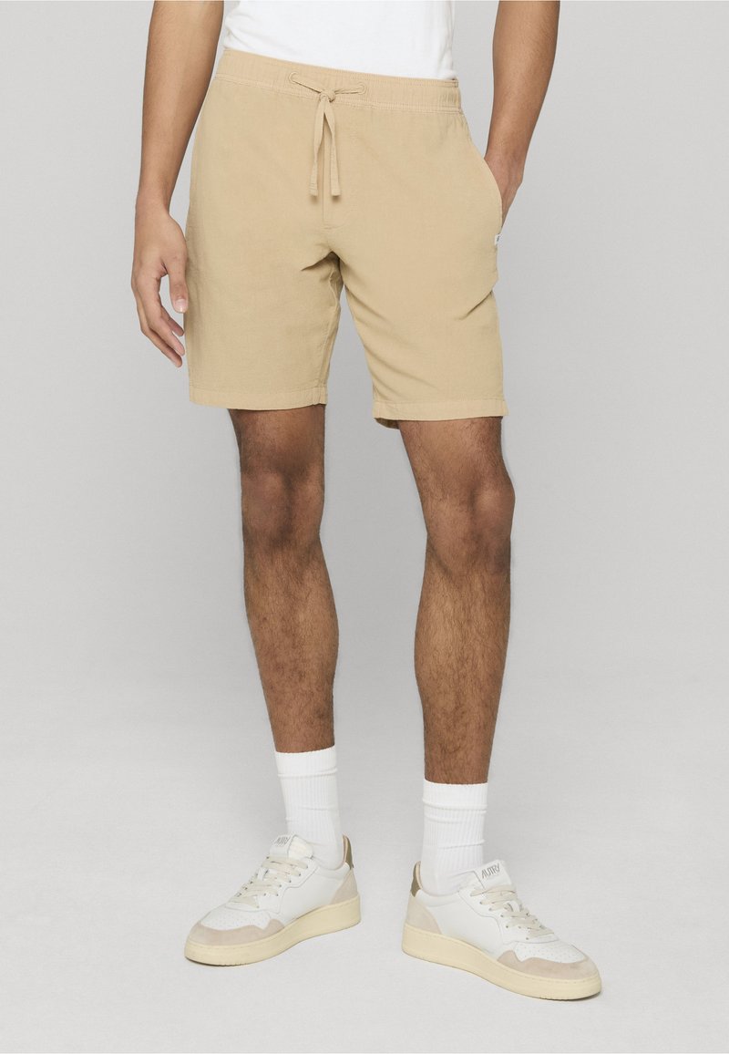 Shorts en coton beige avec une taille élastique, une fermeture à cordon et des poches latérales. Associés à des baskets blanches et des chaussettes blanches côtelées.