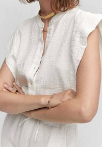 Camicia bianca, strutturata, a maniche corte con dettagli a bottone. Un choker dorato e bracciali completano l'outfit. Design casual con una vestibilità morbida.