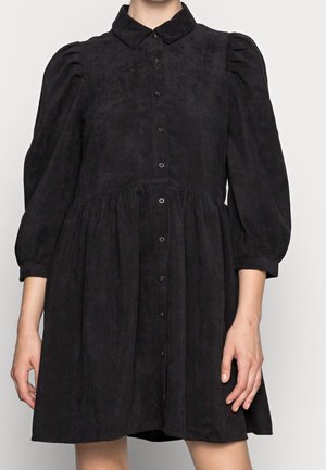 Zwarte corduroy jurk met pofdriekwart mouwen, knoopsluiting aan de voorkant, getailleerde taille en puntige kraag gedragen door een persoon.