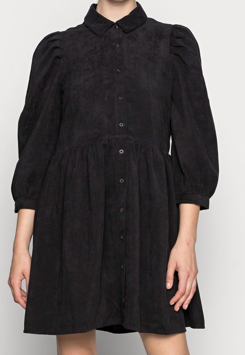 Robe noire en velours côtelé avec manches trois-quarts bouffantes, boutonnée sur le devant, taille cintrée et col pointu portée par une personne.