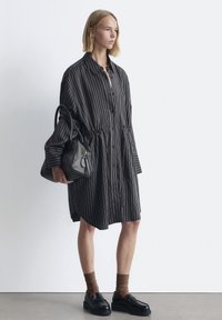 Schwarz-weiß gestreiftes Hemdkleid mit tailliertem Schnitt, langen Ärmeln und Knopfverschluss. Kombiniert mit schwarzen Loafers und einer schwarzen Handtasche.