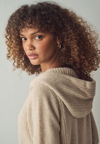 Maglione beige a maglia con cappuccio a coste, caratterizzato da una consistenza morbida e righe verticali. La modella guarda oltre la spalla su uno sfondo chiaro.