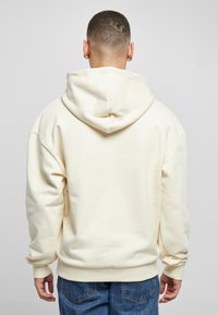 Sudadera color crema con capucha ajustable, corte relajado y puños acanalados. La tela parece suave y lisa. Se muestra la vista trasera.