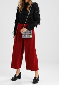 Top nero trasparente con dettagli in frange, pantaloni rossi a gamba larga e borsa a tracolla grigia con decorazioni. Mocassini neri chunky completano il look.