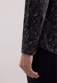 Camicia a maniche lunghe in tessuto scuro con motivo paisley. Presenta un polsino con bottone e una texture liscia. Include dettagli intricati nelle tonalità di grigio e marrone.