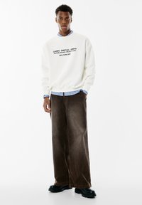 Felpa crema con scritte nere, indossata sopra una camicia a righe blu. Pantaloni in corduroy marrone a gamba larga e stivali neri chunky.