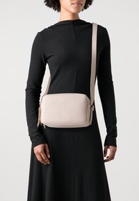 Crossbody bag in soft beige pebbled leather with a rectangular shape, zippered compartments, and a adjustable strap. Subtle brand logo displayed.
