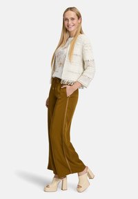 Haakjasje in het wit, versierd met franjes; crèmekleurige blouse met bloemdessin; wijde mosterdkleurige broek; beige sandalen met blokhak.