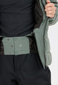 Giubbotto imbottito con un guscio esterno verde chiaro, foderato in nero, chiusura con zip e bottoni a pressione in vita. Tessuto testurizzato e polsini elasticizzati.