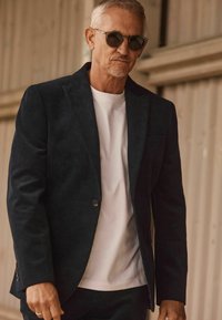 Black corduroy blazer with textured finish, single button closure, worn over a white t-shirt, paired with round black sunglasses.