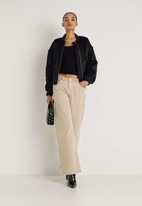 Μαύρο μπουφάν bomber, μαύρο cropped τοπ, μπεζ παντελόνι με φαρδιά μπατζάκια και μαύρα μποτάκια μέχρι τον αστράγαλο. Κρατά μικρή μαύρη τσάντα με λευκές πουά.