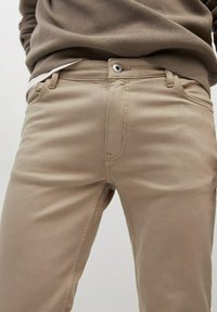 Beżowe bawełniane spodnie o gładkiej fakturze, posiadające pojedynczy guzik i rozporek, ozdobione przeszyciami oraz wyposażone w boczne kieszenie.